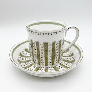 May include: A white teacup and saucer set with a green and brown geometric pattern. The cup has a handle and the saucer has a scalloped edge.