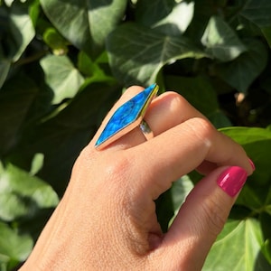 May include: A silver ring featuring a large, elongated diamond-shaped blue stone with a vibrant, iridescent surface. The stone is set in a smooth, polished silver bezel that follows the sharp, angular geometry of the gemstone's unique shape.