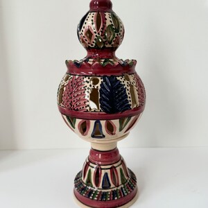 Könnte beinhalten: Ein Kerzenhalter aus Keramik mit einem weißen Sockel und einem roten, grünen und blauen Muster. Der Halter hat eine runde, offene Oberseite und einen dekorativen Sockel.