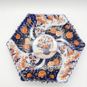 Può includere: Un piatto esagonale in porcellana con un motivo floreale blu e bianco. Il piatto ha un bordo ondulato ed è decorato con accenti dorati.