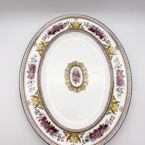 May include: An oval, white ceramic serving platter with a floral design. The rim features a green border, with a decorative pattern of gold, purple, and pink flowers. A smaller floral design is centered on the platter.