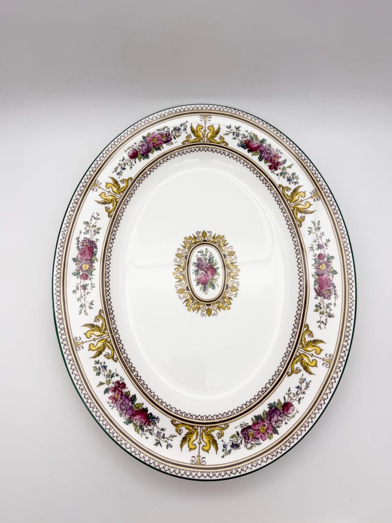 May include: An oval, white ceramic serving platter with a floral design. The rim features a green border, with a decorative pattern of gold, purple, and pink flowers. A smaller floral design is centered on the platter.