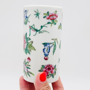 Può includere: Un bicchiere in ceramica bianca decorato con motivi floreali e insetti colorati. Il design include fiori rosa, rossi, blu e verdi, insieme a una mantide religiosa verde. Il bicchiere è cilindrico e tenuto in mano.