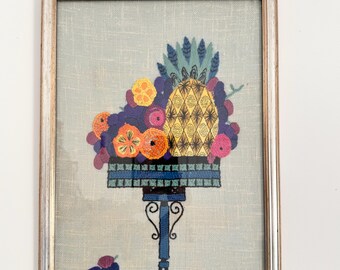 May include: A framed embroidery of a pineapple and other fruits in a blue and green basket on a blue stand. The fruits are yellow, orange, red, and purple.