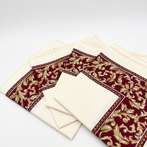 May include: A set of folded cream-coloured napkins featuring a wide border with a deep red background and a repeating gold-toned acanthus leaf scroll pattern. The centre of each napkin has a subtle, light-coloured dotted texture throughout the fabric.