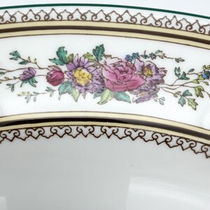 May include: Close-up of a white ceramic plate with a floral design. The plate features a border with a brown and gold pattern, and a green edge. The floral design includes pink, purple, and yellow flowers with green leaves.