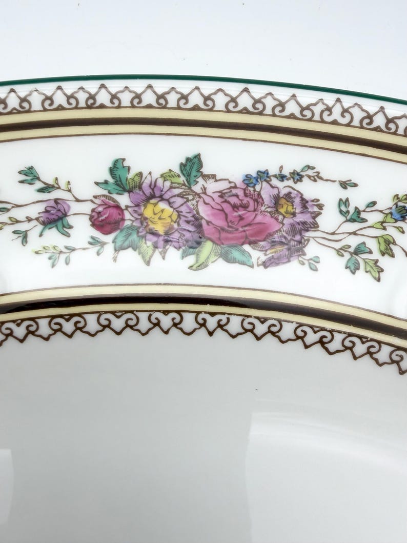 May include: Close-up of a white ceramic plate with a floral design. The plate features a border with a brown and gold pattern, and a green edge. The floral design includes pink, purple, and yellow flowers with green leaves.