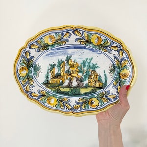 Pu&ograve; includere: Piatto da portata ovale in ceramica con bordo floreale blu e bianco e una scena centrale di un edificio giallo e marrone con alberi verdi e cielo blu.
