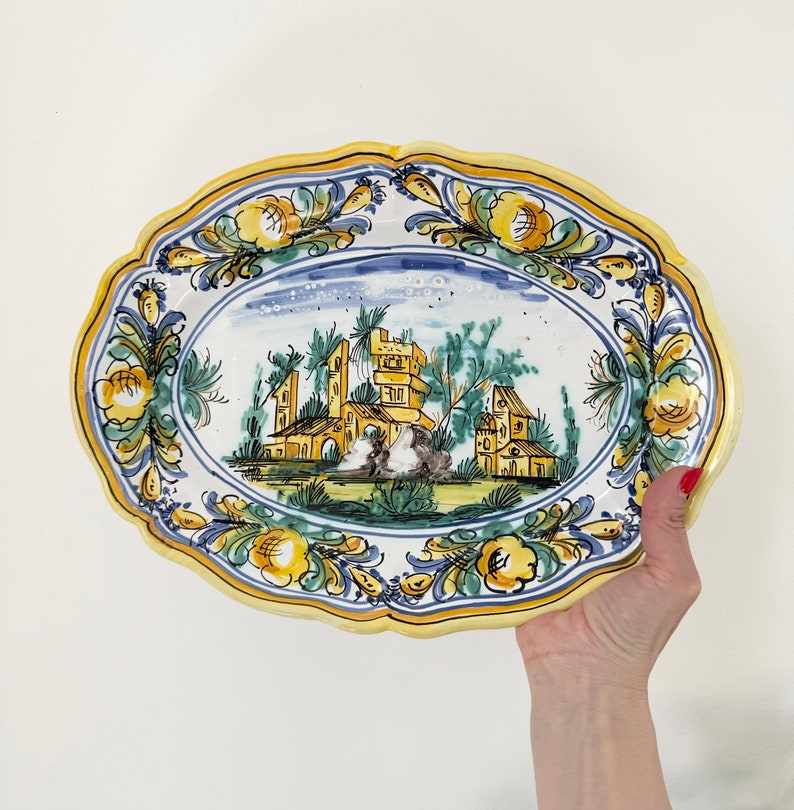 Pu&ograve; includere: Piatto da portata ovale in ceramica con bordo floreale blu e bianco e una scena centrale di un edificio giallo e marrone con alberi verdi e cielo blu.