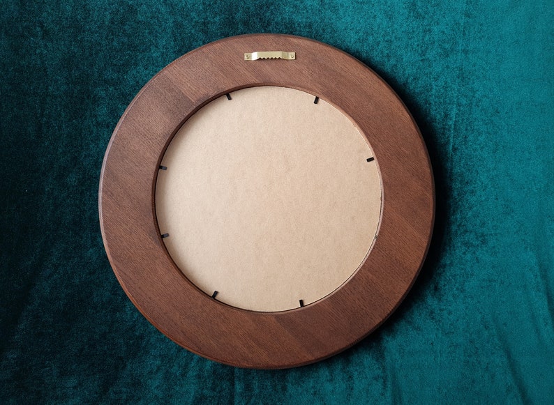 May include: A round wooden frame with a brown finish. The frame has a cardboard backing and a metal hanging hook on the back.
