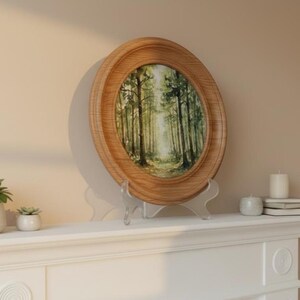 Op de afbeelding: Een ovaalvormige houten lijst met een aquarel van een boslandschap met hoge bomen en licht dat door het bladerdak valt. De lijst staat op een doorzichtige acrylstandaard en toont een natuurlijke houtnerf en een gladde afwerking van het hout.