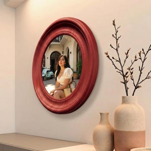 May include: A circular wall mirror featuring a deep red, wood-grain textured frame. The reflective glass surface displays a scene of a person sitting outdoors near a light blue scooter, with architectural details visible in the background reflection.
