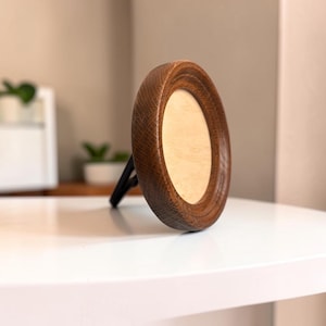 Op de afbeelding: Een ronde houten fotolijst met een donkerbruine gebeitste afwerking en een zichtbare houtnerf. De lijst heeft een lichtbeige, gladde, ronde inzet in het midden en wordt ondersteund door een zwarte standaard die aan de achterkant is bevestigd.