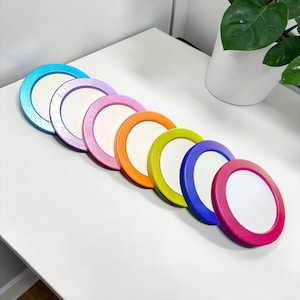 Pode incluir: Uma coleção de sete bases para copos redondas e coloridas. Cada base tem um centro branco e uma borda colorida em tons de azul, roxo, rosa, laranja, verde e magenta. As bases estão dispostas em diagonal sobre uma superfície branca.