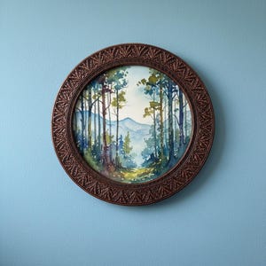 Op de afbeelding: Een rond wandkunstwerk met een aquarel landschapsschilderij van een bos met hoge bomen en verre blauwe bergen. Het kunstwerk is gevat in een donkerbruine, sierlijke, uitgesneden ronde lijst met een gedetailleerd bladerpatroon.