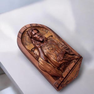 Op de afbeelding: Een houten wandplaquette met een gegraveerd beeld van Jezus Christus met een gouden hart op zijn borst. De plaquette is gemaakt van donker hout met een gouden rand.