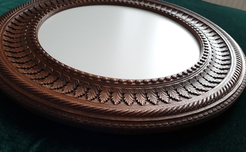 May include: A round, wooden mirror with a detailed, carved frame. The frame features a rope-like design and a row of leaves around the edge.