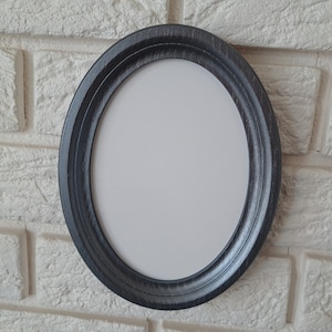 Puede incluir: Marco de fotos ovalado gris oscuro con interior blanco, montado en una pared de ladrillos blancos. El marco tiene un aspecto texturizado y ligeramente desgastado. El marco está vacío, listo para una foto o una obra de arte.