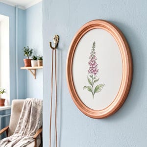 May include: An oval-shaped copper-coloured frame containing a botanical illustration of a pink foxglove flower with green leaves on a white background. The frame is mounted on a light blue wall next to a brass-coloured hook with a thin cord hanging down.