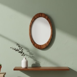 Puede incluir: Espejo ovalado con marco de madera colgado en una pared verde claro. El marco tiene un acabado de vetas de madera natural. Debajo, una estantería de madera marrón con un jarrón blanco y vegetación.