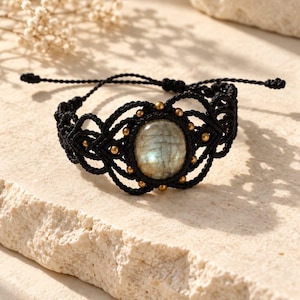 Könnte beinhalten: Ein schwarzes Makramee-Armband mit einem ovalen Labradorit-Edelstein in der Mitte. Der Stein ist von kleinen metallisch goldenen Perlen und komplizierten geknoteten Kordelmustern umgeben. Das Armband verfügt über verstellbare Schiebekordeln.