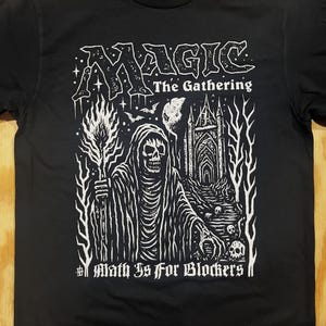 May include: Black t-shirt featuring a white graphic for "Magic: The Gathering." The design includes a hooded figure with a skull face, a burning torch, a gothic cathedral, and the text "Math Is For Blockers."