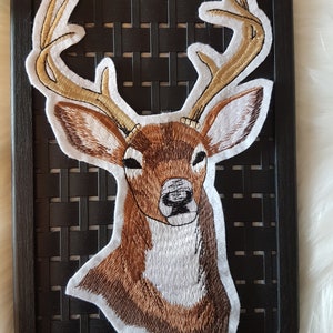 Könnte beinhalten: Gestickter Aufnäher mit dem Kopf eines braunen und weißen Rehs mit großen Geweih. Das Reh schaut nach vorne, der Kopf ist leicht nach rechts geneigt. Das Geweih ist hellbraun und weiß umrandet.