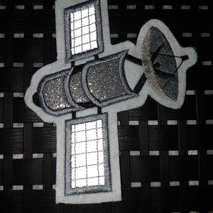 Könnte beinhalten: Ein grauer und weißer Satelliten-Patch mit glitzerndem Design. Der Satellit hat eine große Parabolantenne und zwei Solarpaneele.