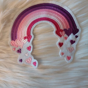 Könnte beinhalten: Ein regenbogenfarbener Filz-Patch mit einer Reihe von rosa, roten und violetten Herzen, die auf jeder Seite gestickt sind.