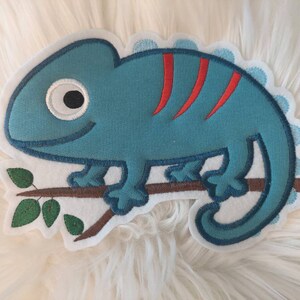 Könnte beinhalten: Ein gestickter Aufnäher mit einem blauen Chamäleon auf einem Ast mit grünen Blättern. Das Chamäleon hat rote Streifen und ein weißes Auge.