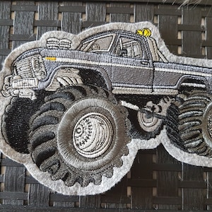 Könnte beinhalten: Ein grauer gestickter Patch mit einem Monstertruck mit großen Reifen. Der Truck hat eine schwarze Kabine und einen grauen Körper. Die Reifen sind schwarz mit weißem Profil.