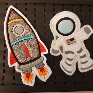 Könnte beinhalten: Gestickte Aufnäher mit einer Rakete und einem Astronauten. Die Rakete hat einen grauen Körper, rote Flossen und orangefarbene Flammen. Der Astronaut ist weiß mit grauen Akzenten und einem blauen Visier. Ideal für Weltraumprojekte.