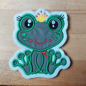 Könnte beinhalten: Ein grüner und grauer Frosch, der eine gelbe Krone mit roten Akzenten trägt. Der Frosch hat große schwarze Augen und ein breites Lächeln. Der Frosch sitzt auf einem weißen Hintergrund.