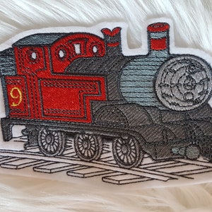Könnte beinhalten: Gestickter Aufnäher eines roten und grauen Spielzeug-Lokomotiv-Motors mit schwarzen Rädern auf weißem Hintergrund. Der Motor hat die Nummer "9" an der Seite.