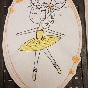 Könnte beinhalten: Ovaler Aufnäher aus weißem Filz mit einer Ballerina in einem gelben Tutu, umrandet mit schwarzem Faden. Das Design umfasst einen gelben Haargummi und Ballettschuhe. Der Rand ist mit orangefarbenem Faden bestickt und mit orangefarbenen Herzen verziert.