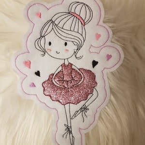 Könnte beinhalten: Eine rosa und weiße Filzapplikation einer Ballerina mit einem rosa Glitzer-Tutu und schwarzen Haaren. Die Ballerina ist von rosa Herzen umgeben.