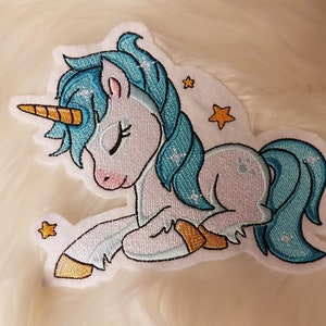 Könnte beinhalten: Ein gestickter Aufnäher mit einem blauen und weißen Einhorn mit geschlossenem Auge, das mit dem Kopf zur Seite liegend. Das Einhorn hat ein goldenes Horn und eine blaue Mähne und einen blauen Schwanz. Im Hintergrund befinden sich kleine gelbe Sterne.