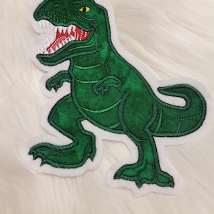 Könnte beinhalten: Grüner gestickter Dinosaurier-Patch mit weißem Umriss. Der Dinosaurier steht auf seinen Hinterbeinen, mit offenem Maul und sichtbaren Zähnen. Der Dinosaurier hat ein gelbes Auge.