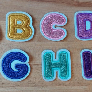 Könnte beinhalten: Zehn Buchstaben des Alphabets, A bis J, aus Stoff mit Glitzerdetails. Jeder Buchstabe hat eine kontrastierende Umrandung und ist vor einem weißen Hintergrund platziert. Die Buchstaben sind in verschiedenen Farben, darunter Silber, Gold, Rosa, Lila und Rot, auf einer Holzoberfläche.