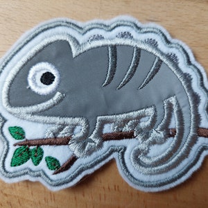 Könnte beinhalten: Ein grauer gestickter Aufnäher mit einem Chamäleon, das auf einem Ast mit grünen Blättern sitzt. Das Chamäleon hat ein großes Auge und ein gestreiftes Muster auf dem Rücken.