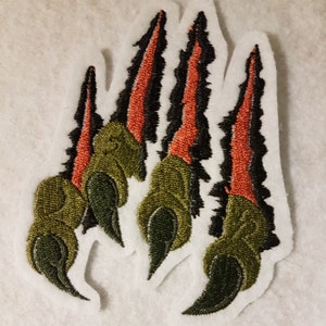 Könnte beinhalten: Gestickter Aufnäher mit einem grünen und schwarzen Krallendesign. Die Krallen sind spitz und scharf, mit roten Akzenten. Der Aufnäher ist perfekt, um Jacken, Taschen oder anderen Accessoires einen Hauch von kantigem Stil zu verleihen.