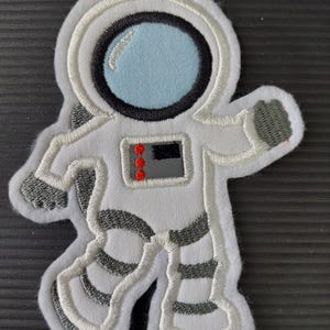 Könnte beinhalten: Weißer gestickter Aufnäher eines Astronauten mit schwarzem Helm und blauem Visier. Der Astronaut trägt einen weißen Raumanzug mit schwarzen und silbernen Streifen.
