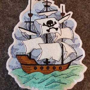 Könnte beinhalten: Gestickter Aufnäher eines Piratenschiffs, das auf blauem Wasser mit einer Totenkopf- und Knochenflagge segelt. Das Schiff hat braune Segel und einen weißen Rumpf mit schwarzen Akzenten.