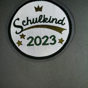 Könnte beinhalten: Ein runder weißer Patch mit grünem Rand. Der Patch hat eine goldene Krone, den Text "Schulkind 2023" in grüner und goldener Schrift und zwei goldene Sterne.