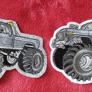 Könnte beinhalten: Zwei gestickte Patches mit einem grauen Monstertruck mit großen Reifen. Die Patches befinden sich auf einem roten Stoffhintergrund.