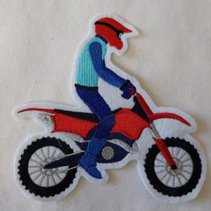 Könnte beinhalten: Gestickter Aufnäher mit einem Motocross-Fahrer auf einem rot-schwarzen Motorrad. Der Fahrer trägt einen roten Helm, ein blaues Hemd und eine blaue Hose. Der Aufnäher hat einen weißen Rand und ist für Kleidung oder Accessoires bestimmt.