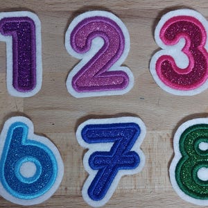Könnte beinhalten: Zehn glitzernde Zahlenaufnäher, von Null bis Neun, mit weißem Rand. Die Zahlen sind in verschiedenen Farben wie Rosa, Lila, Rot, Blau, Grün und Gold gehalten. Geeignet für Bastelarbeiten oder Lernzwecke.