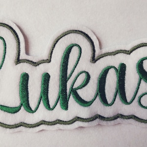 Könnte beinhalten: Ein weißer Filz-Patch mit einem grünen gestickten Namen "Lukas" in einer kursivschrift.