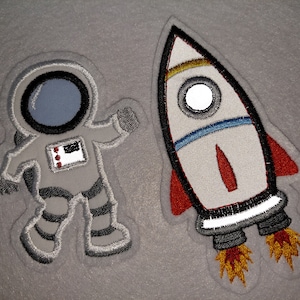 Könnte beinhalten: Eine graue Filzapplikation eines Astronauten in einem weißen Raumanzug mit schwarzem Helm und einem rot-weiß-blauen Aufnäher auf der Brust. Neben dem Astronauten befindet sich eine graue Filzapplikation einer Rakete mit roten, weißen, blauen und gelben Details.
