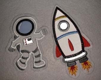 May include: A gray felt appliqué of an astronaut in a white spacesuit with a black helmet and a red, white, and blue patch on the chest. Next to the astronaut is a gray felt appliqué of a rocket ship with red, white, blue, and yellow details.